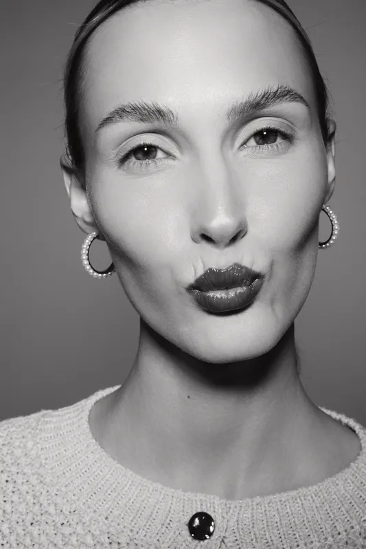 Caucasian woman with fair skin makes a kiss face, wearing hoop earrings and a knit sweater in black and white portrait.