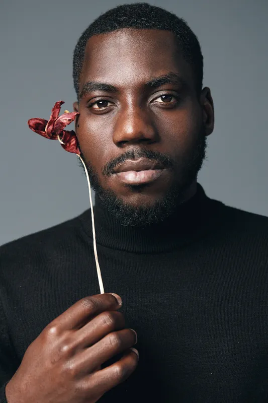African man with dark skin holding dried flower near face in creative conceptual visual style. Portrait of thoughtful black male model with beard wearing black turtleneck on gray background.