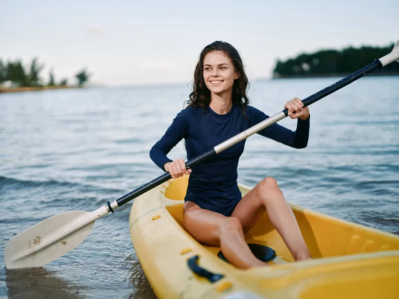 Female kayaker paddling a bright yellow kayak on calm water, wearing a dark blue long sleeve top, smiling confidently, enjoying a sunny outdoor paddling session.