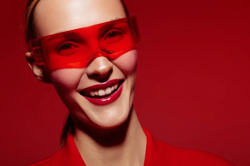 Smiling woman wearing red transparent glasses and red outfit poses on red background. Concept of fashion, style, beauty, and modern accessories with vibrant color theme.