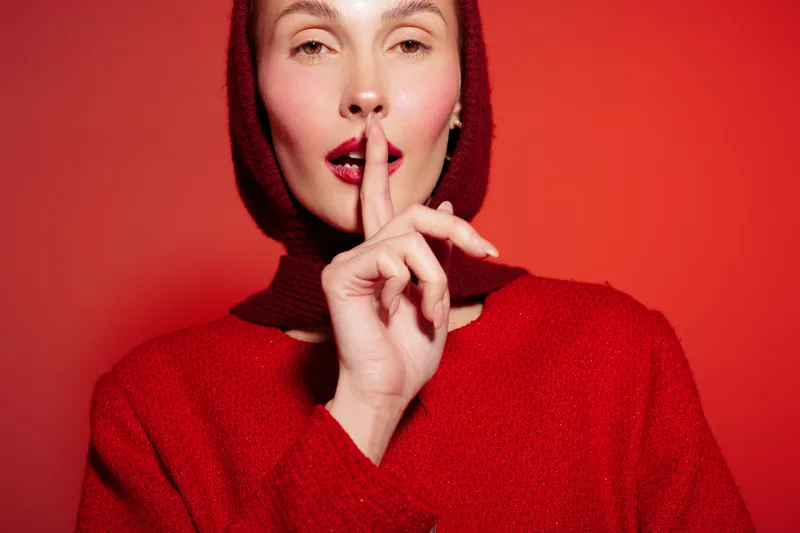 Woman in red sweater and hood makes silence gesture with finger on lips against red background. Concept of secrecy, quiet, and calm mood in studio portrait photography.