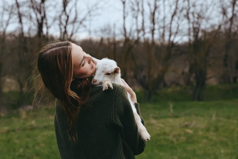 A woman outdoors holds a small goat kid close, expressing affection in a cozy lifestyle moment. Tender bond between human and animal in a park setting with natural light and open green space.