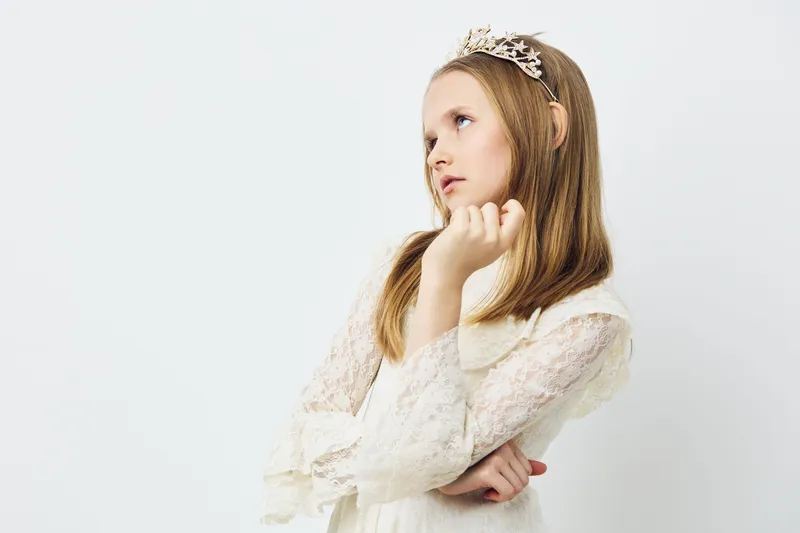 A young girl with long hair wearing a delicate white dress and tiara, expressing a contemplative look against a light background