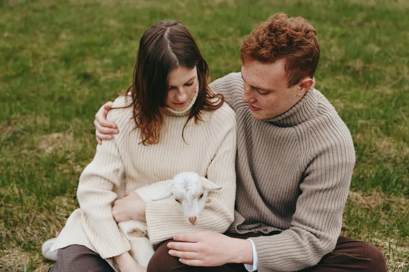 Young couple in cozy sweaters sits on grass gently holding a small white baby goat in their arms enjoying a peaceful moment outdoors in soft natural light.