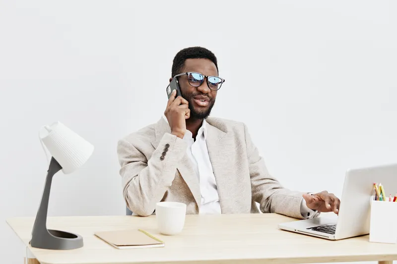 Confident young African American man wearing glasses, beige blazer, and white shirt talking on mobile phone while sitting at a modern desk in bright office environment. People lifestyle concept, profe