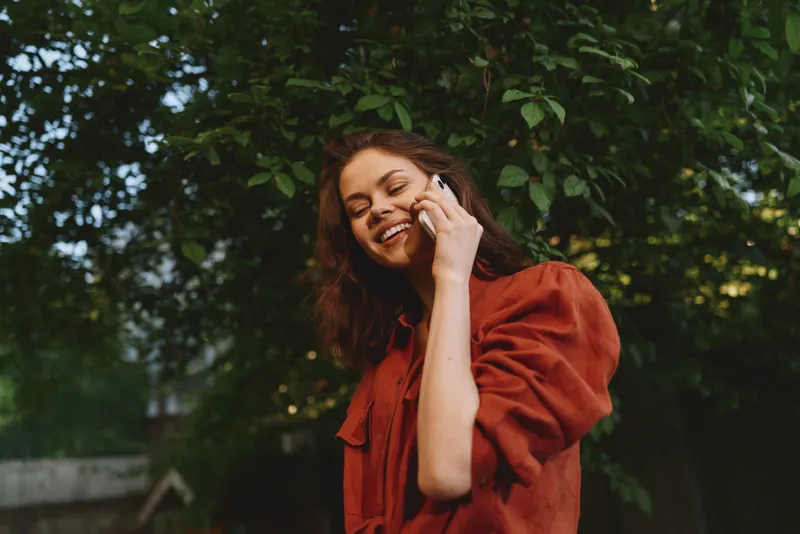Happy woman enjoys a sunny outdoor moment, talking on phone, casual red blouse, natural greenery background, lifestyle portrait highlighting connection and joy