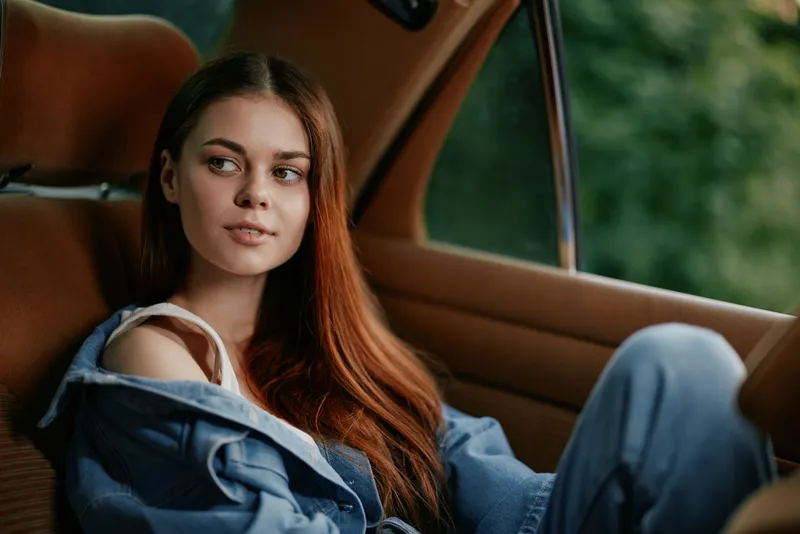 Young woman with long hair in casual outfit, sitting inside a car, evoking calmness and contemplation, surrounded by nature and greenery.