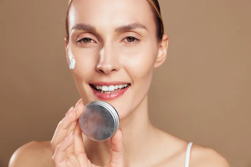 Woman in closeup applies skincare cream to face, beauty routine with antiaging focus, smiling and holding a cosmetic container, smooth skin, bright studio light, concept of personal care and