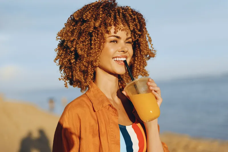 Young woman with curly hair enjoys a refreshing orange juice drink by the seaside on a sunny day while wearing a striped top and an orange shirt, smiling happily.