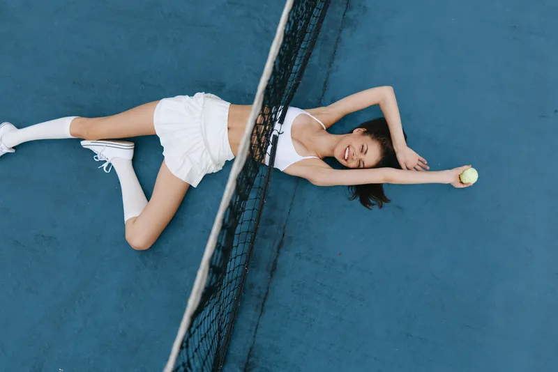 Young woman lying on a tennis court, showcasing a joyful expression while holding a tennis ball, wearing a sporty outfit and white sneakers