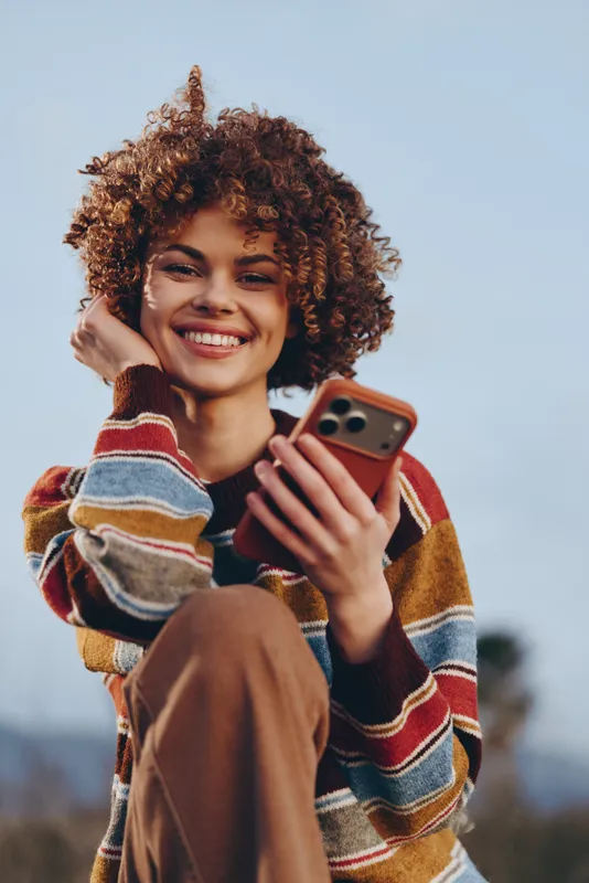 Woman lifestyle, smiling woman with curly hair, holding smartphone near face outdoors, relaxed casual portrait, warm sweater and autumn colors, soft blue sky background, modern communication moment