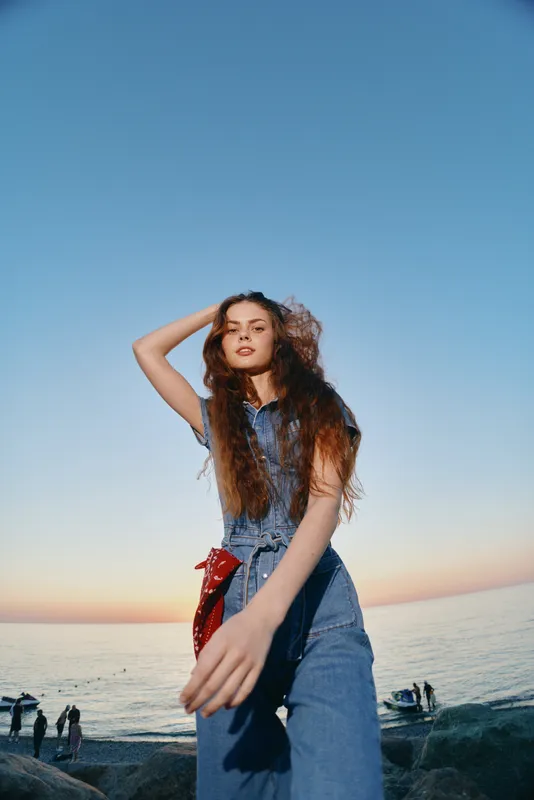 lifestyle woman in modern boho-western denim street style stands confidently by the sea during sunset, showcasing casual fashion with a film color mood