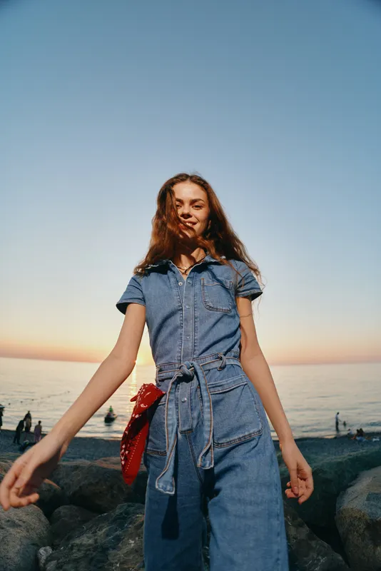 young woman wearing denim jumpsuit and red bandana enjoys sunset on rocky shore with calm sea background, casual style and relaxed summer mood