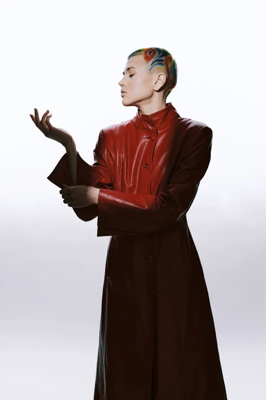 woman with colorful hair wearing a red leather coat posing elegantly against a white background, showcasing modern fashion and bold style in a studio setting.