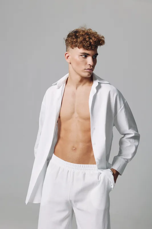 Young man with curly hair wearing white open shirt and pants posing with hand in pocket against plain background. Casual style, confident look and modern fashion portrait.
