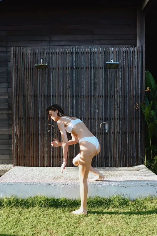Casual woman bikini outdoors, mid motion by wooden wall and shower, sunlit scene, fresh breeze, vibrant green garden, relaxed posture, lifestyle moment, natural colors, candid outdoor fun, movement,