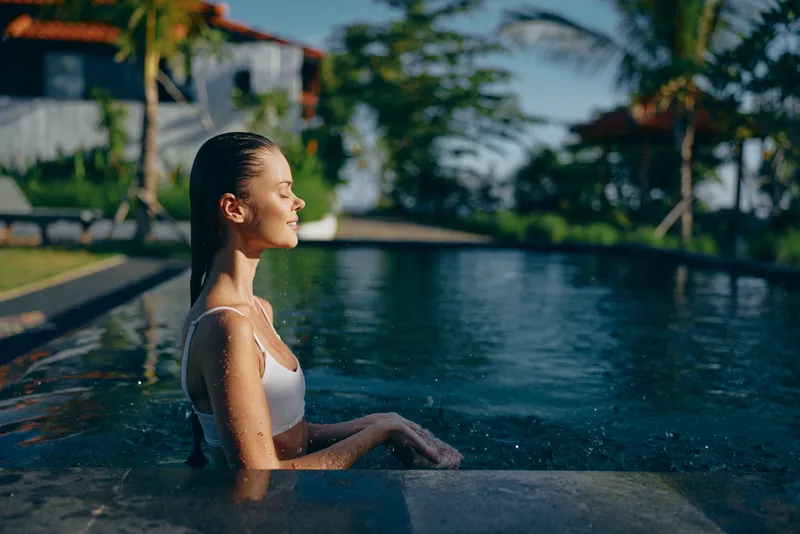 Calm woman in an outdoor pool, profile toward the sun, serene moment by crystal water, lush resort scenery, warm natural tones, relaxed poolside leisure and vacation vibe