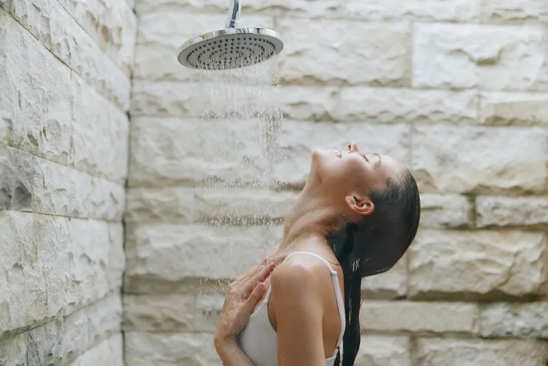 Woman enjoying outdoor shower with water cascading on her face, relaxed and refreshing moment, natural stone wall background, wellness and self-care theme.