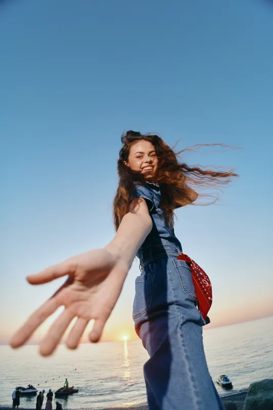 Lifestyle woman wearing modern boho-western denim street style reaching out with sunlit ocean background, film color effect enhancing warm and vibrant atmosphere.