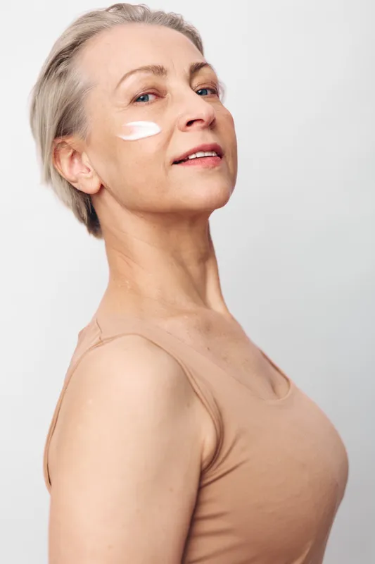Mature woman with short grey hair applies white face cream on cheek. She wears beige top and poses confidently against a plain light background. Concept of skincare, aging, and self-care.