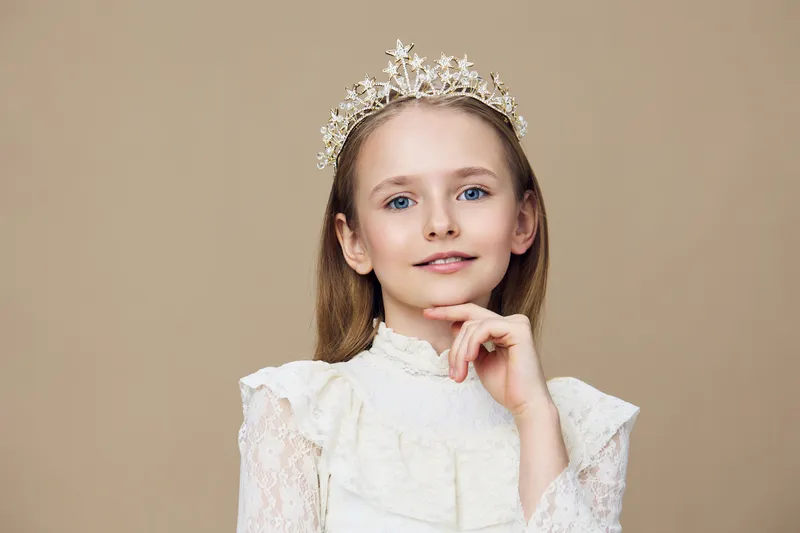 A young girl with a crown smiles confidently against a beige background, wearing an elegant white dress that exudes grace and innocence, perfect for childhood celebrations