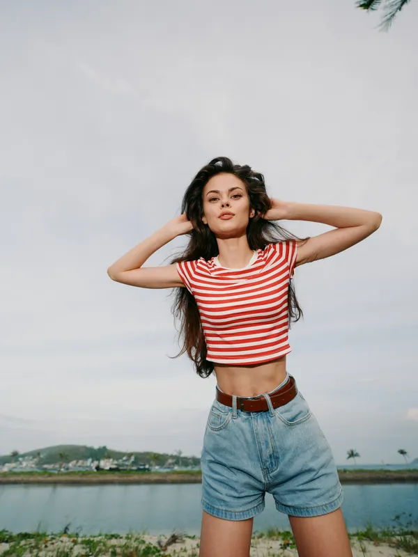 A stylish young woman poses outdoors in a red striped crop top and high waist denim shorts, standing with hands in her hair against a bright sky and tranquil lake backdrop.