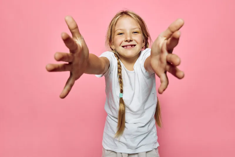 Happy smiling girl with braids wearing white t-shirt reaching out with open hands on pink background.