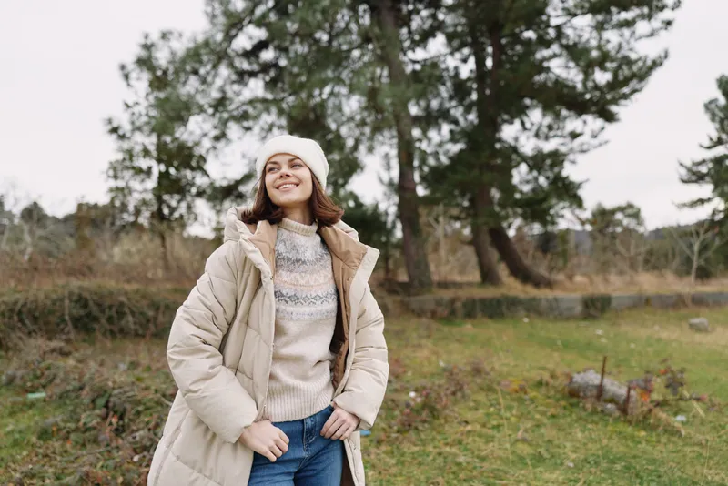 Woman in a warm coat and knit sweater standing outdoors in a park, smiling under a beanie; autumn scenery with trees in the background, casual fashion and relaxed mood.