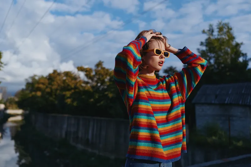 young woman in her early twenties wearing a colorful striped sweater and retro sunglasses, hands on head, leaning by a rural concrete fence under cloudy sky for travel lifestyle.