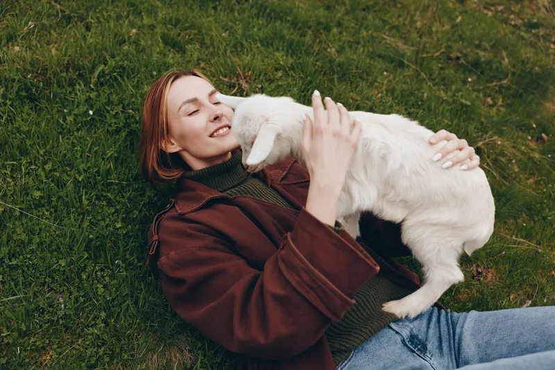 Cheerful woman outdoors, lying on grass, caressing a white goat kid, wearing a brown jacket, warm smile, sunlit moment, gentle breeze, natural colors, rustic countryside, peaceful bonding