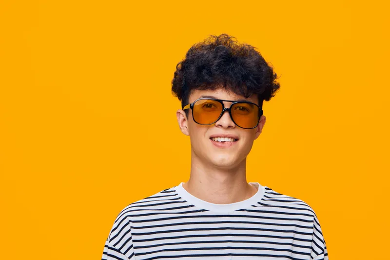 Smiling young man wearing orange sunglasses and striped shirt stands against a bright yellow background, radiating cheerful and trendy vibes with casual hairstyle.