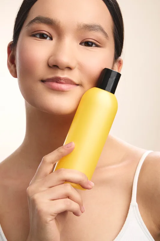 Asian woman smiles softly while showcasing a bright yellow bottle in a clean studio, projecting freshness, simplicity, and a confident skincare vibe.