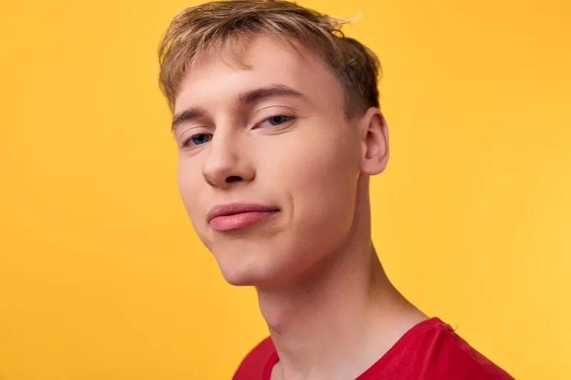 Young man with a bright smile in a red shirt stands against a vibrant yellow background, studio lighting highlights a casual modern fashion look and confident expression.