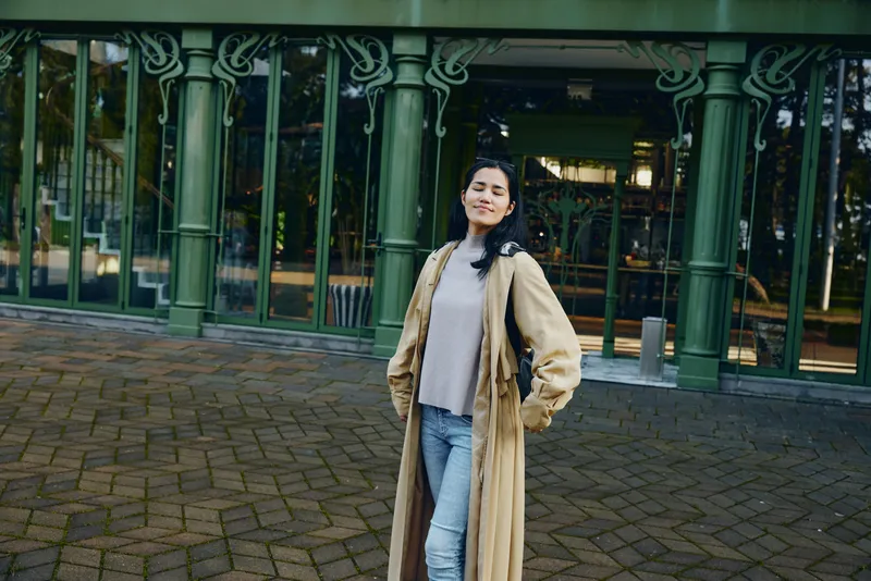Woman wearing a long beige coat and light sweater stands outdoors on a cobbled street in front of a glass storefront, showcasing fashion, confidence, and street style.