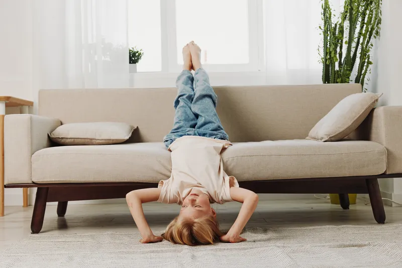 Happy child in casual clothes lies upside down on carpet with legs on couch in bright living room. Concept of playful childhood, leisure, and home comfort for kids.