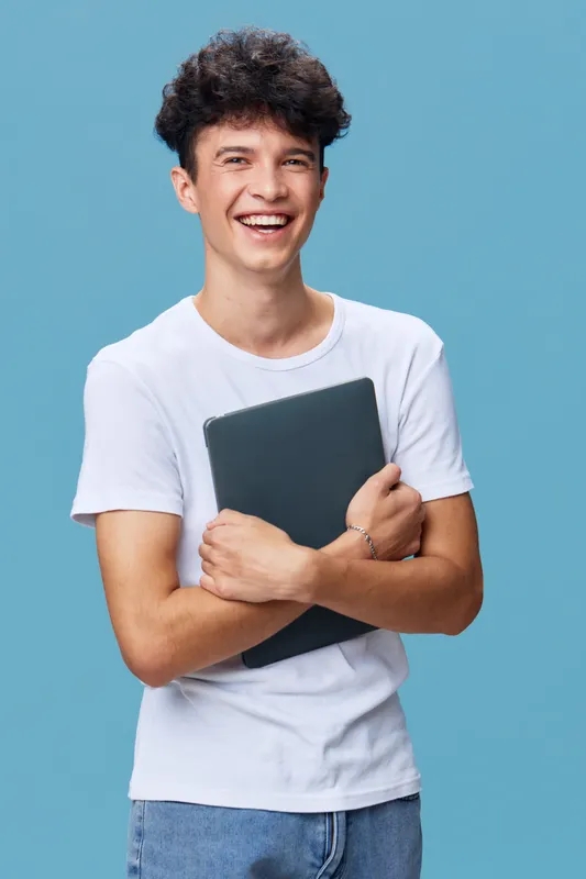 smiling man holding laptop in white casual t-shirt with curly hair on blue background, cheerful young adult expressing happiness and confidence