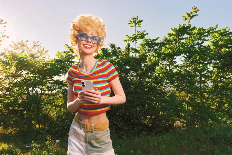 Woman with retro style, striped colorful top and blue sunglasses stands outdoors, holding a phone, bright sunlight, cheerful mood, casual summer vibe, natural greenery in background.