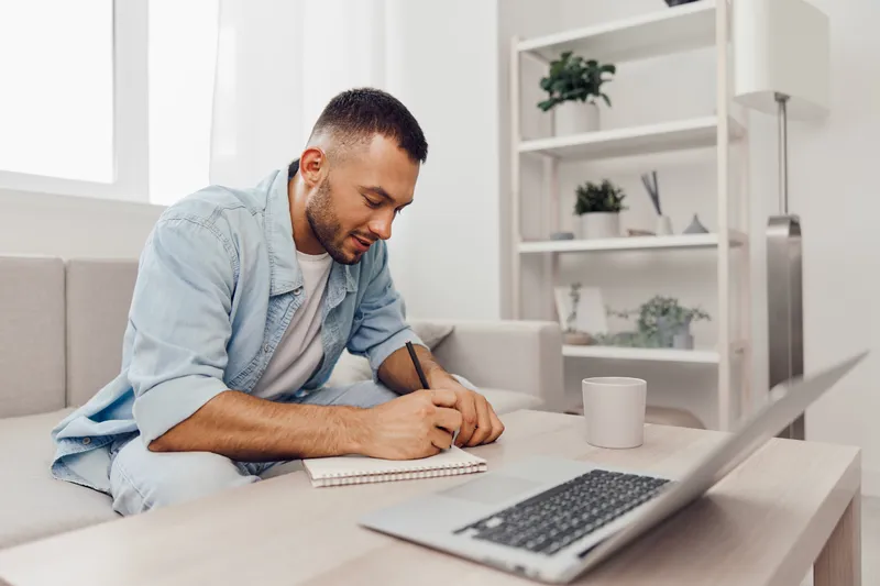 Young man brainstorming ideas at home, sitting on a couch with a notepad and laptop. Casual attire creates a relaxed atmosphere for creativity and productivity.