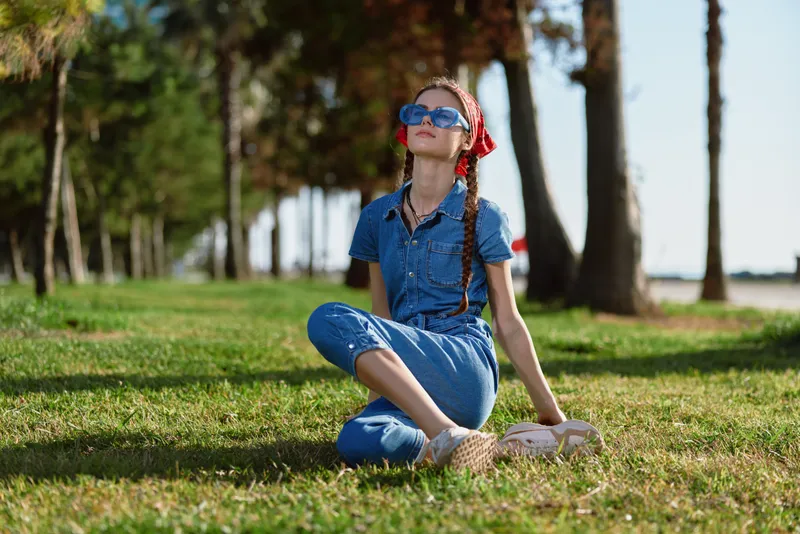 Lifestyle woman in modern boho-western denim street style sitting on grass, wearing sunglasses and a red headband, vibrant film color with natural outdoor light.