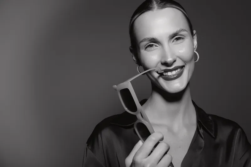 Smiling young Caucasian woman with fair skin holds sunglasses near mouth in studio. Concept of happiness, fashion, and casual lifestyle in black and white.