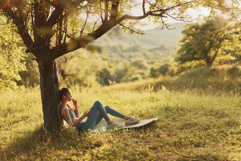 Young woman enjoying a refreshing drink while relaxing under a tree in a vibrant green meadow during golden hour, embodying tranquility and nature