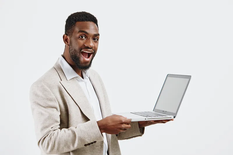 Confident young man holding open laptop computer in studio against plain white background, smiling happily and looking at camera, casual beige blazer, technology device, professionalism, people concep