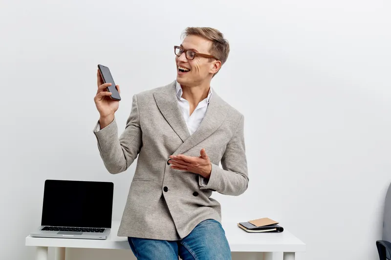 Smiling woman in business casual attire holding smartphone, standing near desk with open laptop and notebooks, cheerful mood, modern office background, technology, communication, happy professional.