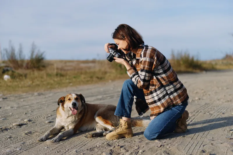 Photographer kneels in a sunny field, capturing a candid portrait of a friendly dog beside a dusty trail, emphasizing light, motion and companionship during outdoor photography.