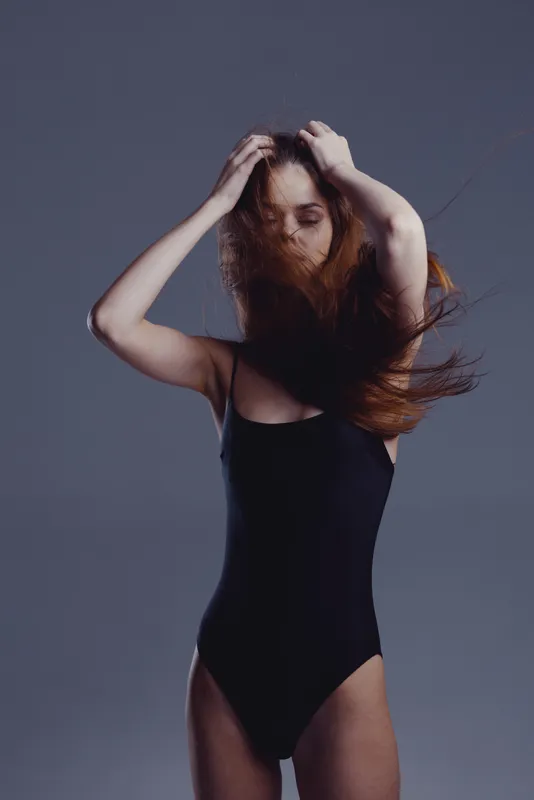 Woman in black bodysuit posing with wind-blown hair against a gray background, expressing calm and movement.