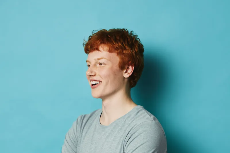 Young man with red hair and freckles smiling and looking to the side wearing gray t shirt on blue background. Casual portrait of male with happy expression and relaxed posture.