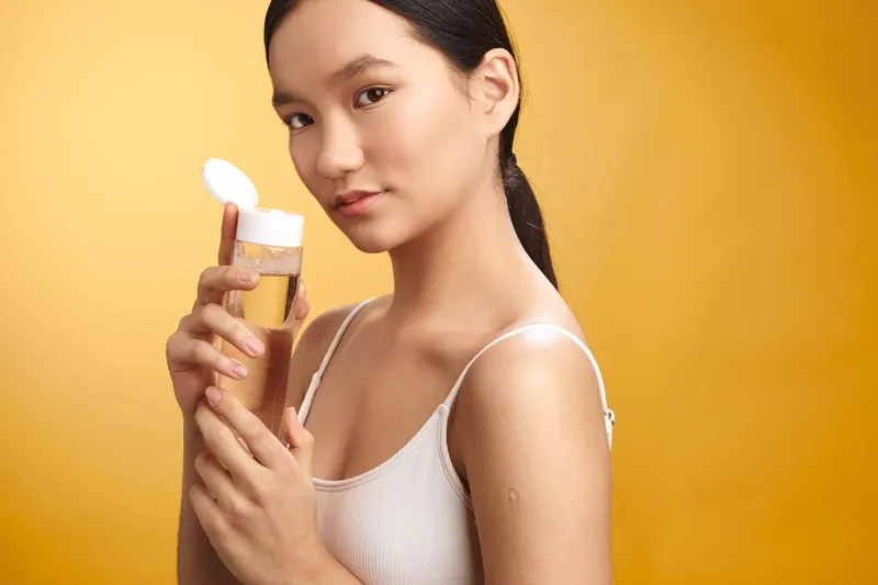 Asian woman applies skincare product in a bright studio, showcasing wellness and beauty. She smiles softly, confident and serene, as vibrant light highlights smooth skin and a calm atmosphere.
