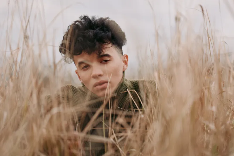 A candid portrait of a young person in a tall grass field, with natural light and a poised gaze that conveys authenticity and credibility in a subtle, unposed moment.