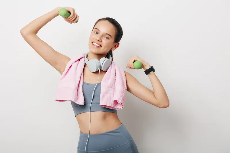 Fitness woman in action with dumbbells and towel, upbeat workout indoors, gray sports bra and leggings, headphones around neck, full of energy and motivation during a dynamic training session.