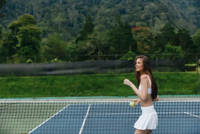 Happy young woman in white sportswear playfully enjoying tennis on a sunny day at an outdoor court with lush green background